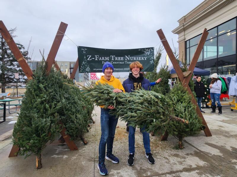 2 people holding trees in front of a Christmas tree lot.